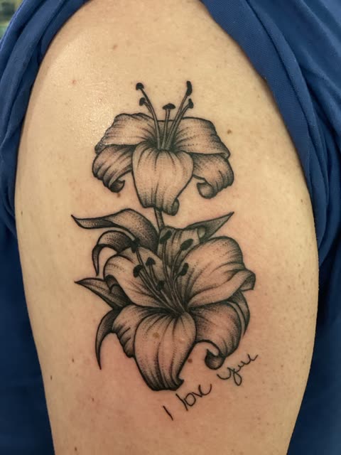 Lilies tattoo with I love you in handwritten script, black whip shading, on an upper arm