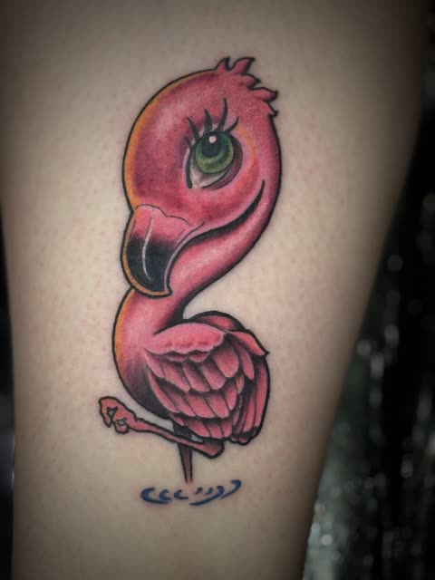 Cartoon pink flamingo tattoo with a green eye, full color, on a lower leg
