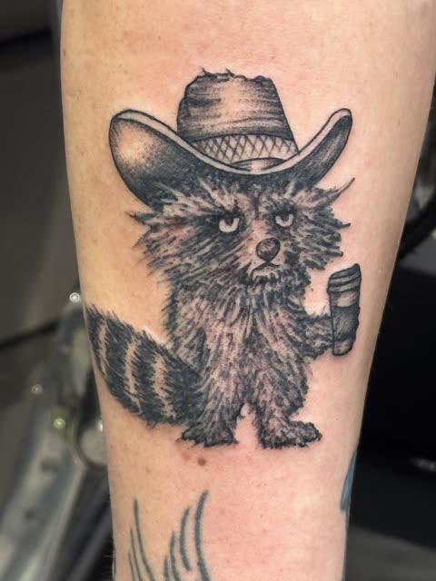 Tattoo of a raccoon in a cowboy hat holding a drink, black whip shading, on a forearm