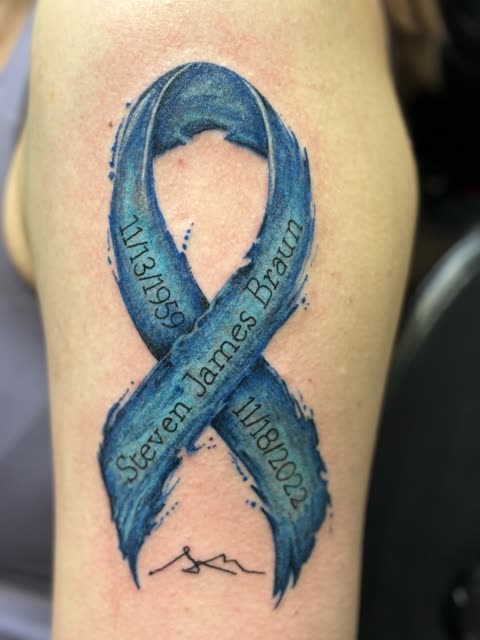 Blue awareness ribbon tattoo in brushed watercolor with a name and dates in script, on an upper arm