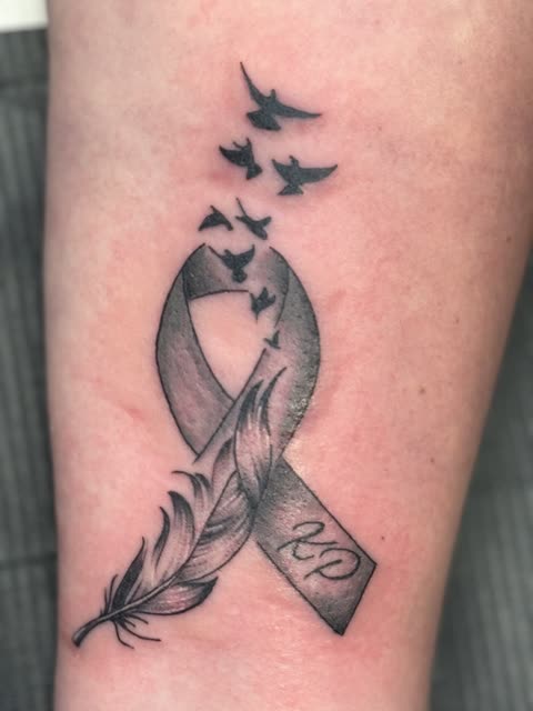 Awareness ribbon tattoo woven with a feather and birds in flight, black shading, on a forearm