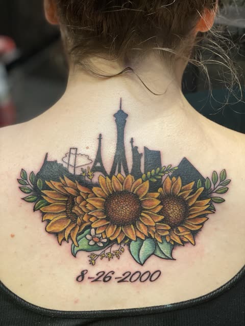 Las Vegas skyline tattoo rising from sunflowers with a date in script, full color, across the upper back