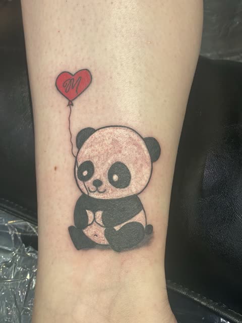 Cartoon panda tattoo with a red heart balloon, black ink with red, on a lower leg