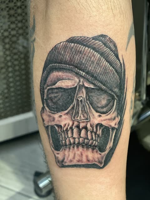 Tattoo of a skull in a knit beanie, black shading, on a forearm