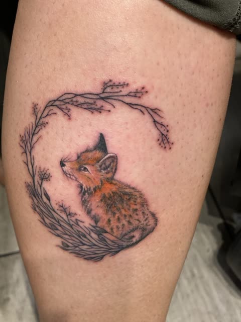 Baby fox tattoo sitting in a wreath of fine line branches, full color, on a leg