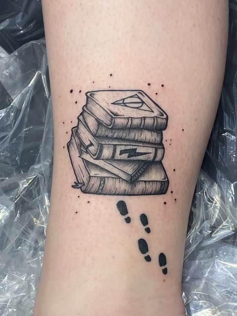 Stack of spellbooks tattoo with footprints walking away, black shading, above the ankle