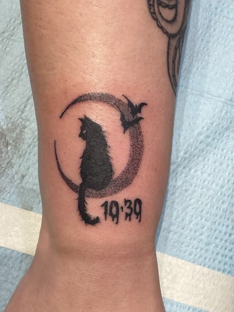 Black cat tattoo sitting in a stippled crescent moon with bats and a time in numbers, above the ankle