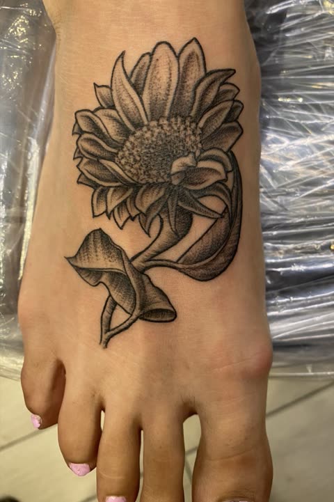 Sunflower tattoo with curling leaves, black stipple shading, on the top of a foot