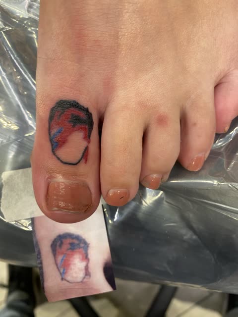 Portrait outline tattoo with a red and blue lightning bolt across the face, on a big toe