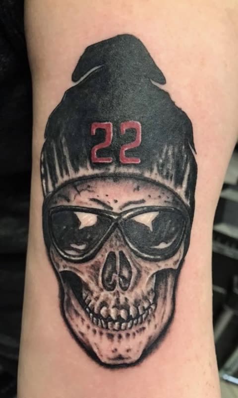 Skull tattoo in a beanie marked 22 wearing sunglasses, black shading with red, on an upper arm