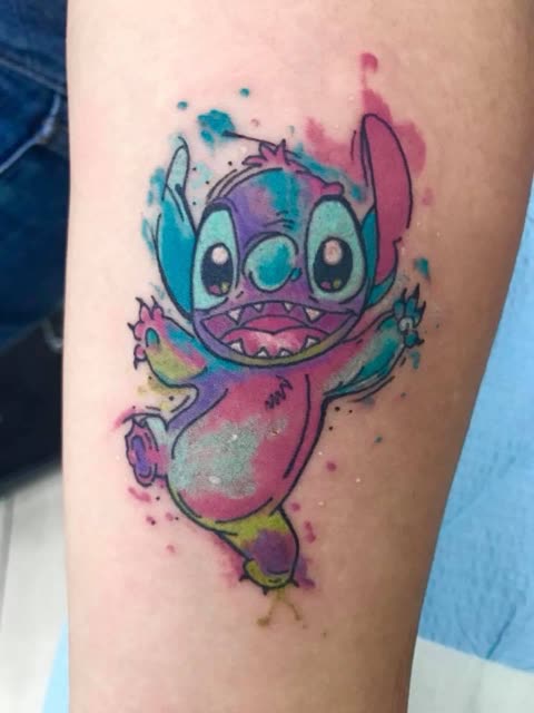 Smiling cartoon alien tattoo in pink, purple and blue watercolor on a forearm